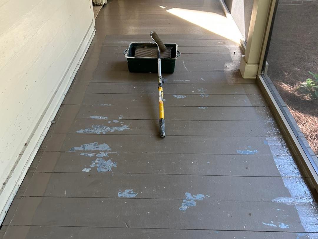 A floor being painted with a paint roller and tray, outdoors. Brown paint on wooden planks.