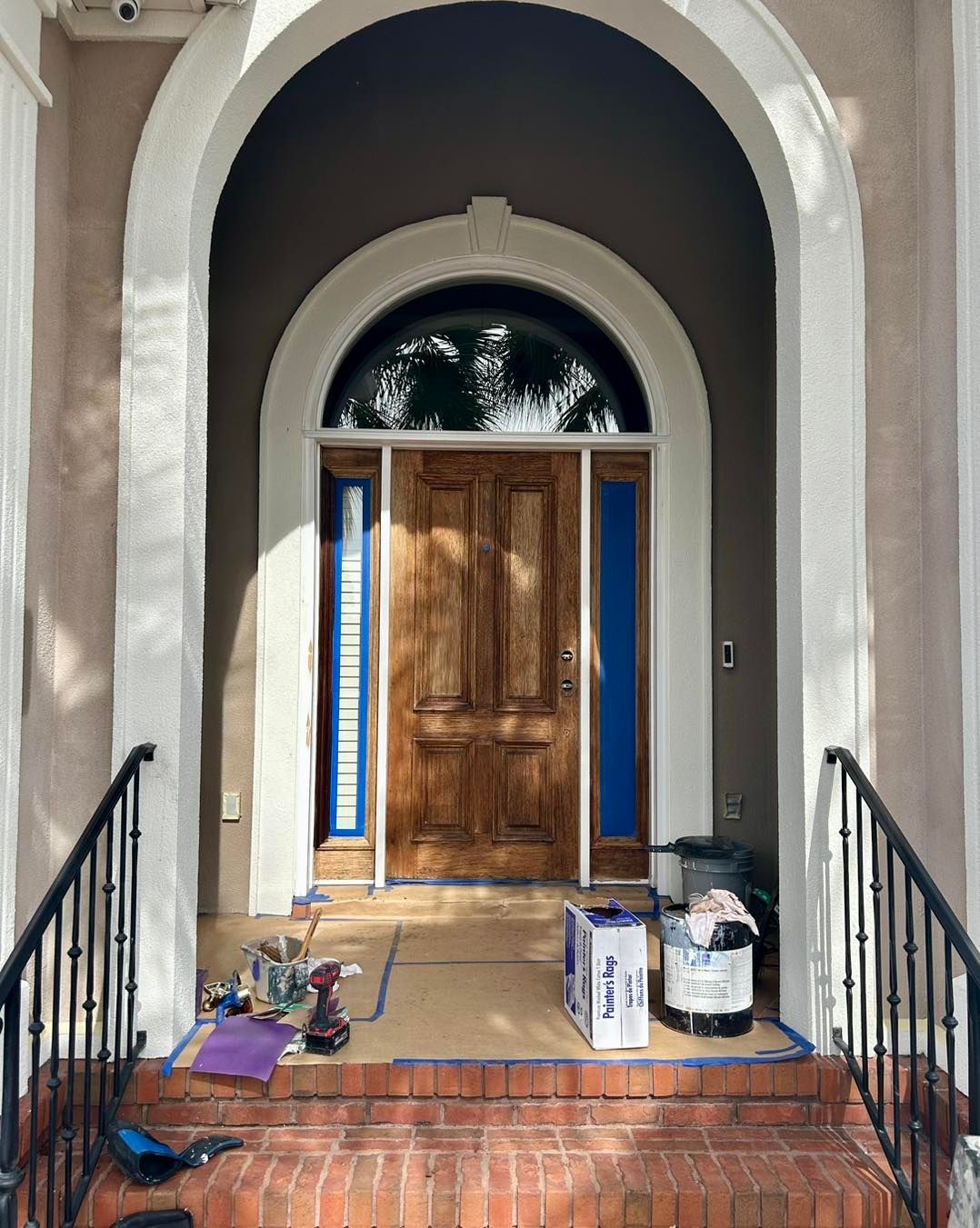 Exterior view of a front door with an arched entryway. The door is stained, and blue tape protects the edges.