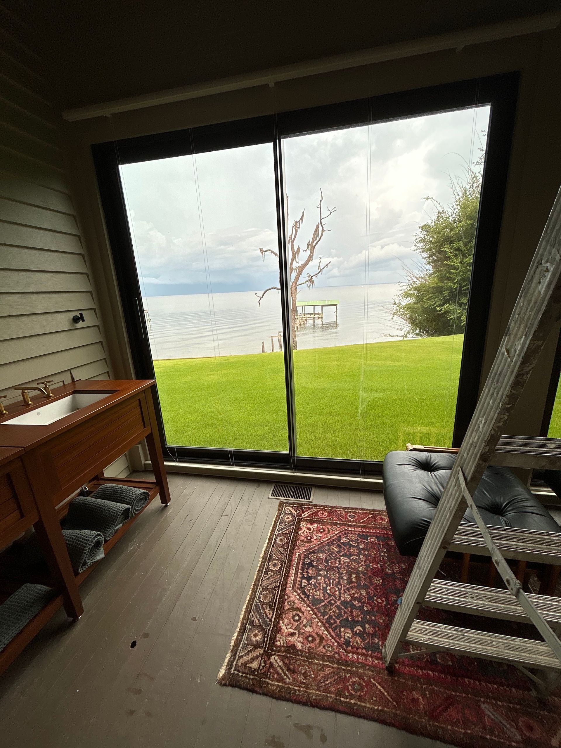 A bathroom with a view of the lake. Wooden vanity, rug, ladder, and a large window.