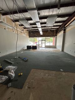 Interior of a gutted retail space with exposed ceiling, damaged flooring, and entrance door visible.