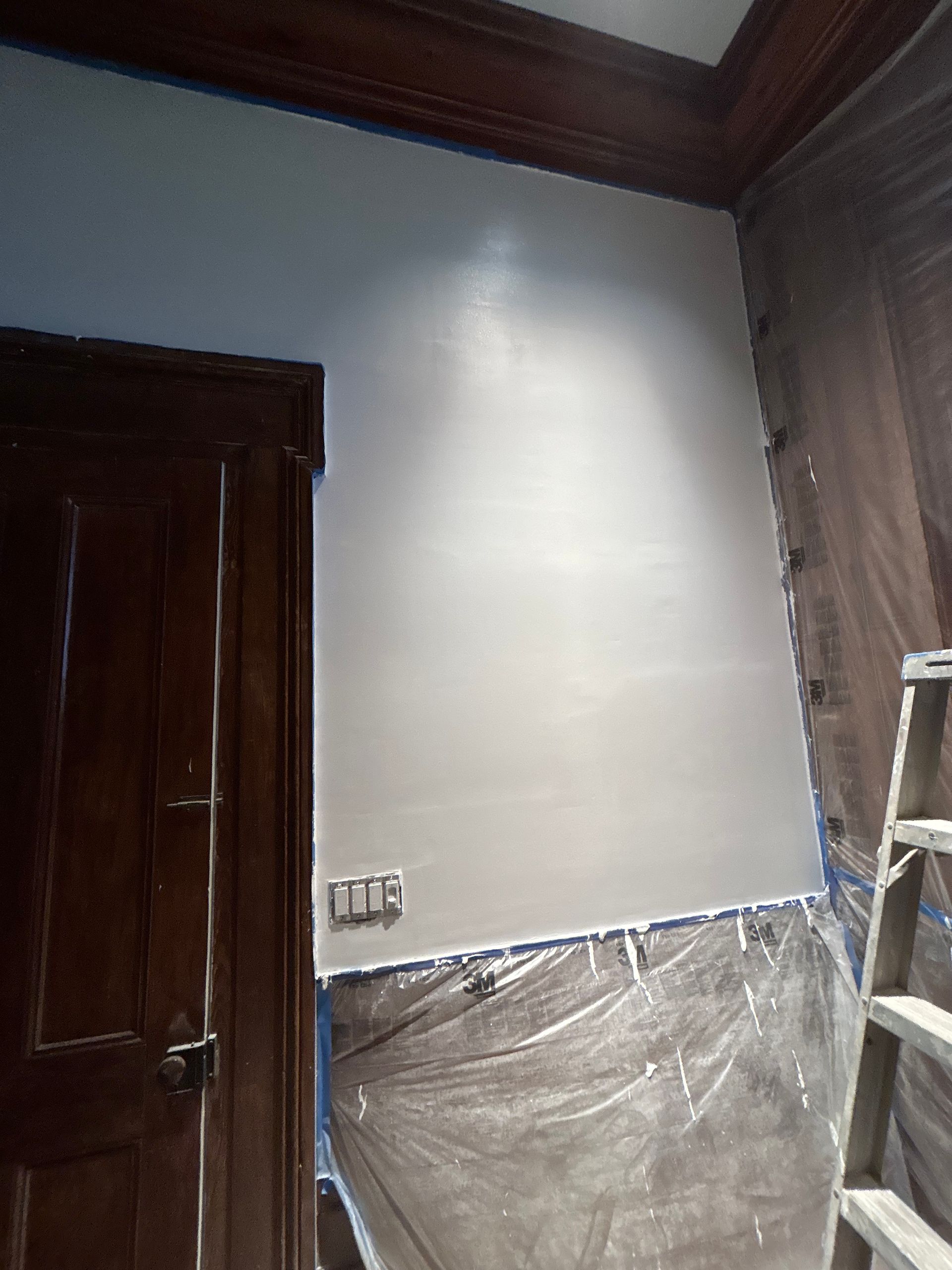 A room with a freshly painted light gray wall; dark wooden door, trim, and ladder visible.