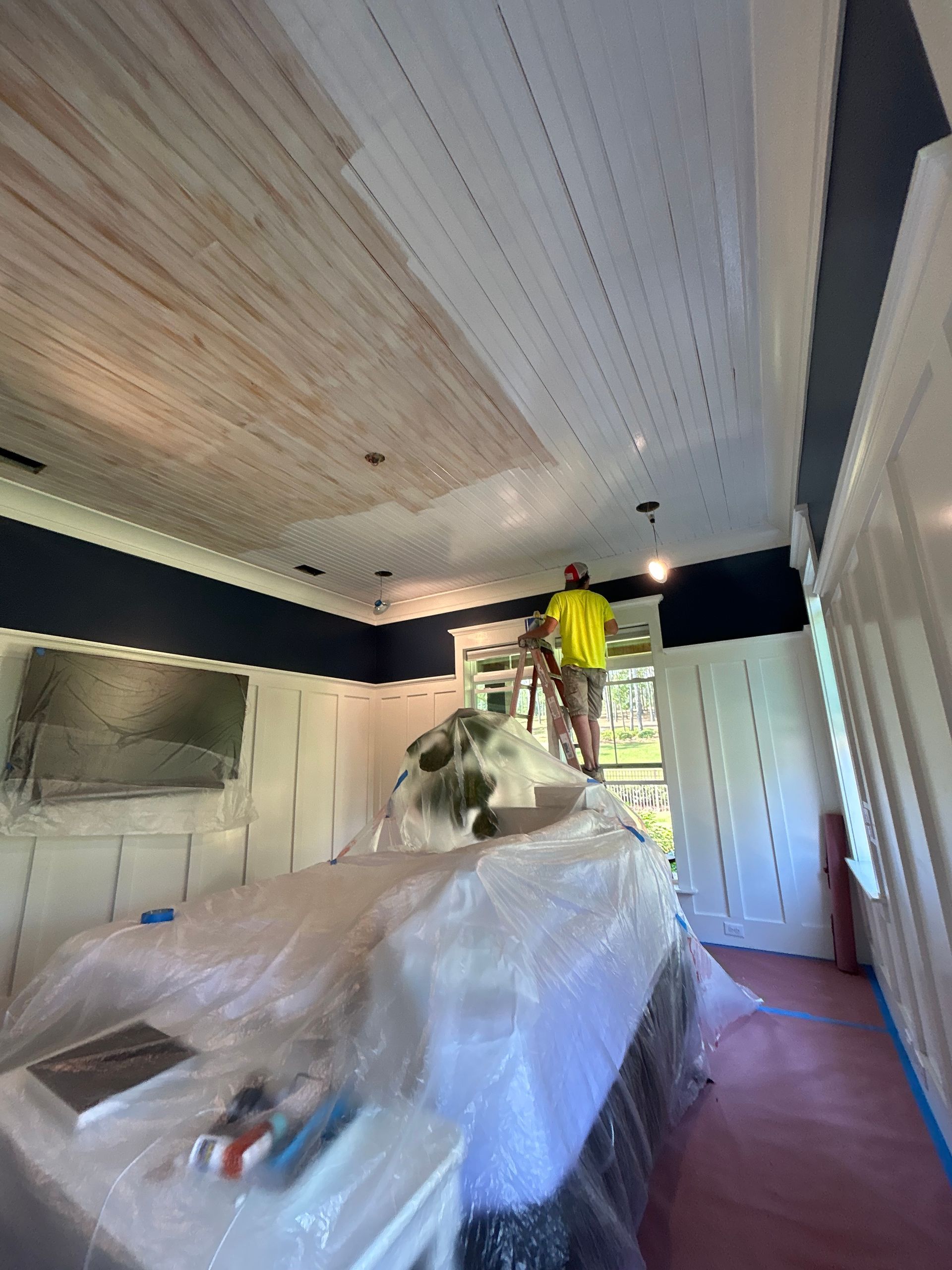 Person painting a white ceiling with wood paneling. Interior room with blue accents, covered furniture.