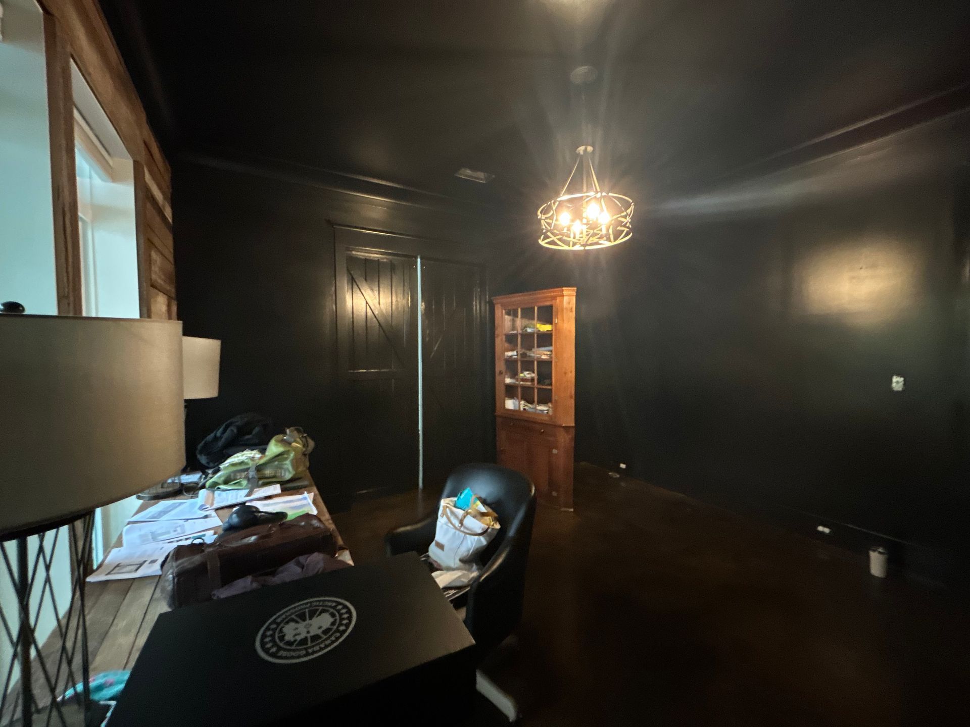 Black-walled room with dark floor, desk with items, ornate light fixture, wooden cabinet, and open door.