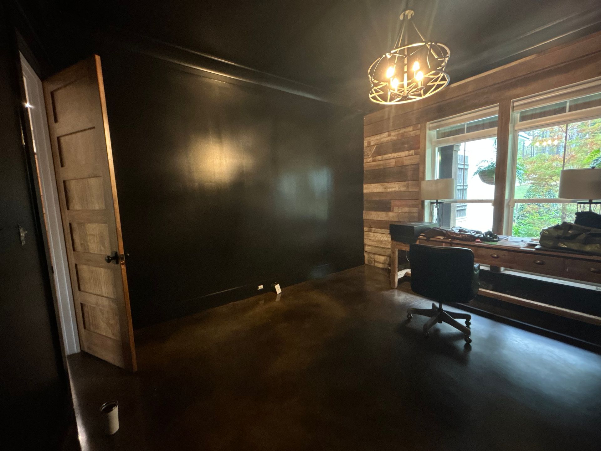 Room with dark walls, wooden door, and a desk by a window; a chandelier hangs from the ceiling.