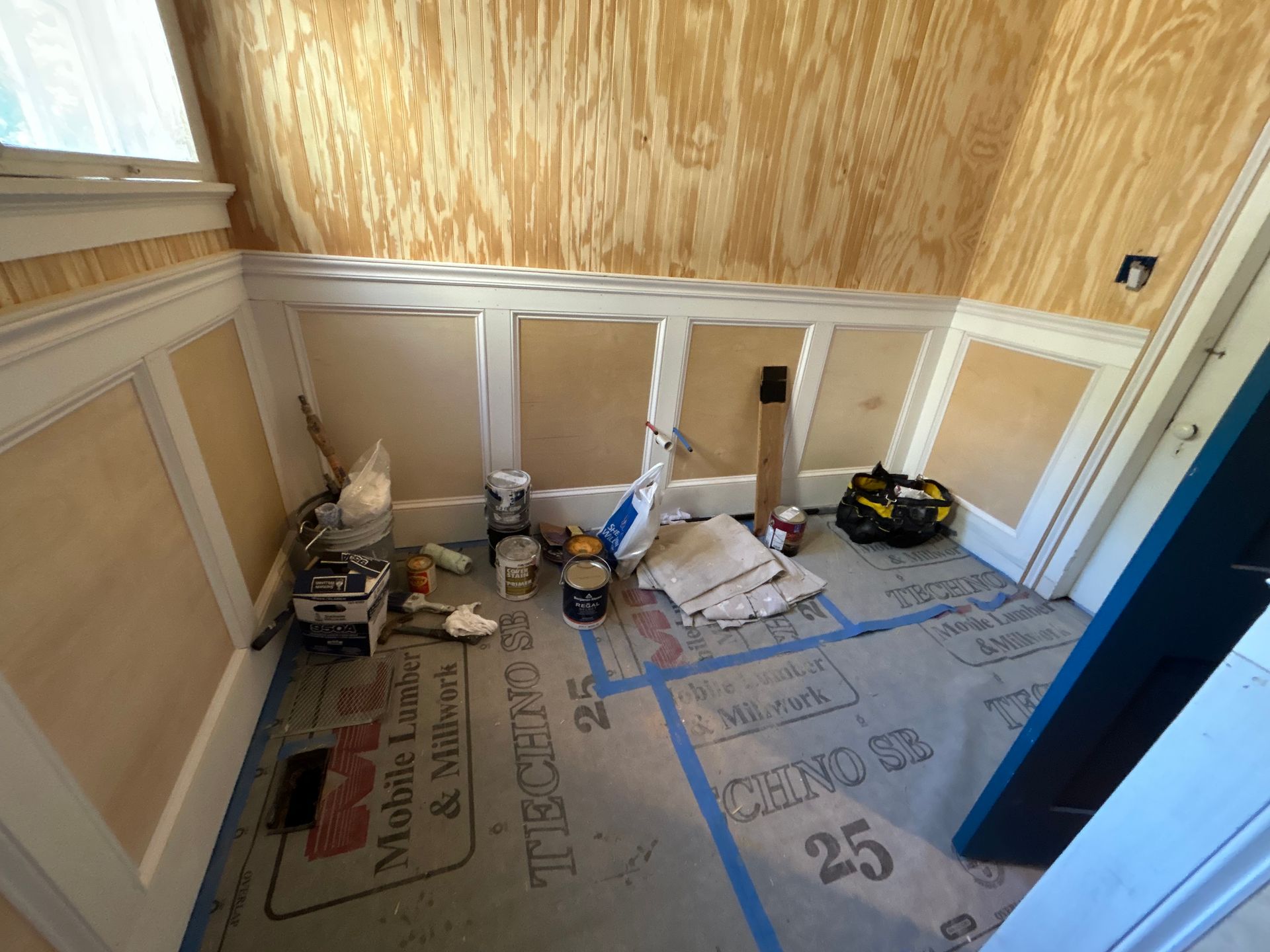 Room under construction with plywood walls and wainscoting, tools and materials scattered on the floor.