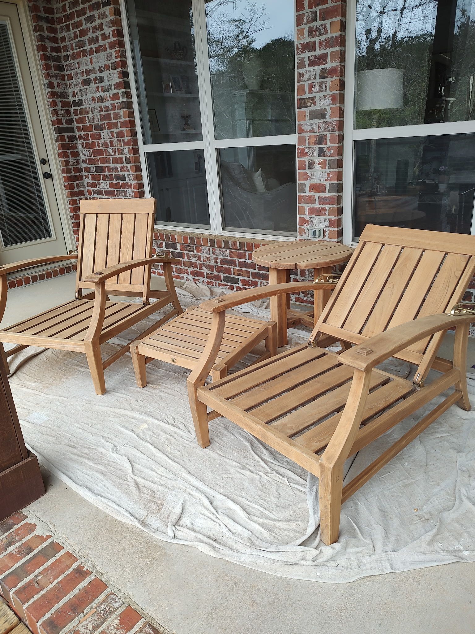 Outdoor teak chairs on a porch, including a lounge chair, chair with ottoman, and another chair, on a cloudy day.
