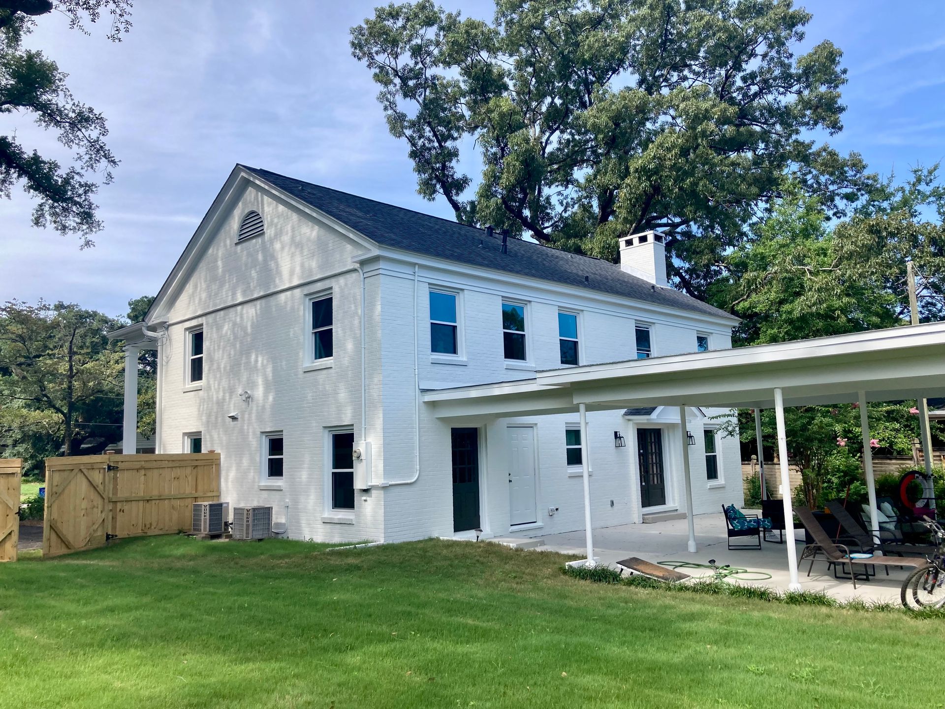 White two-story house with dark roof and attached carport, set on green lawn with trees and blue sky.
