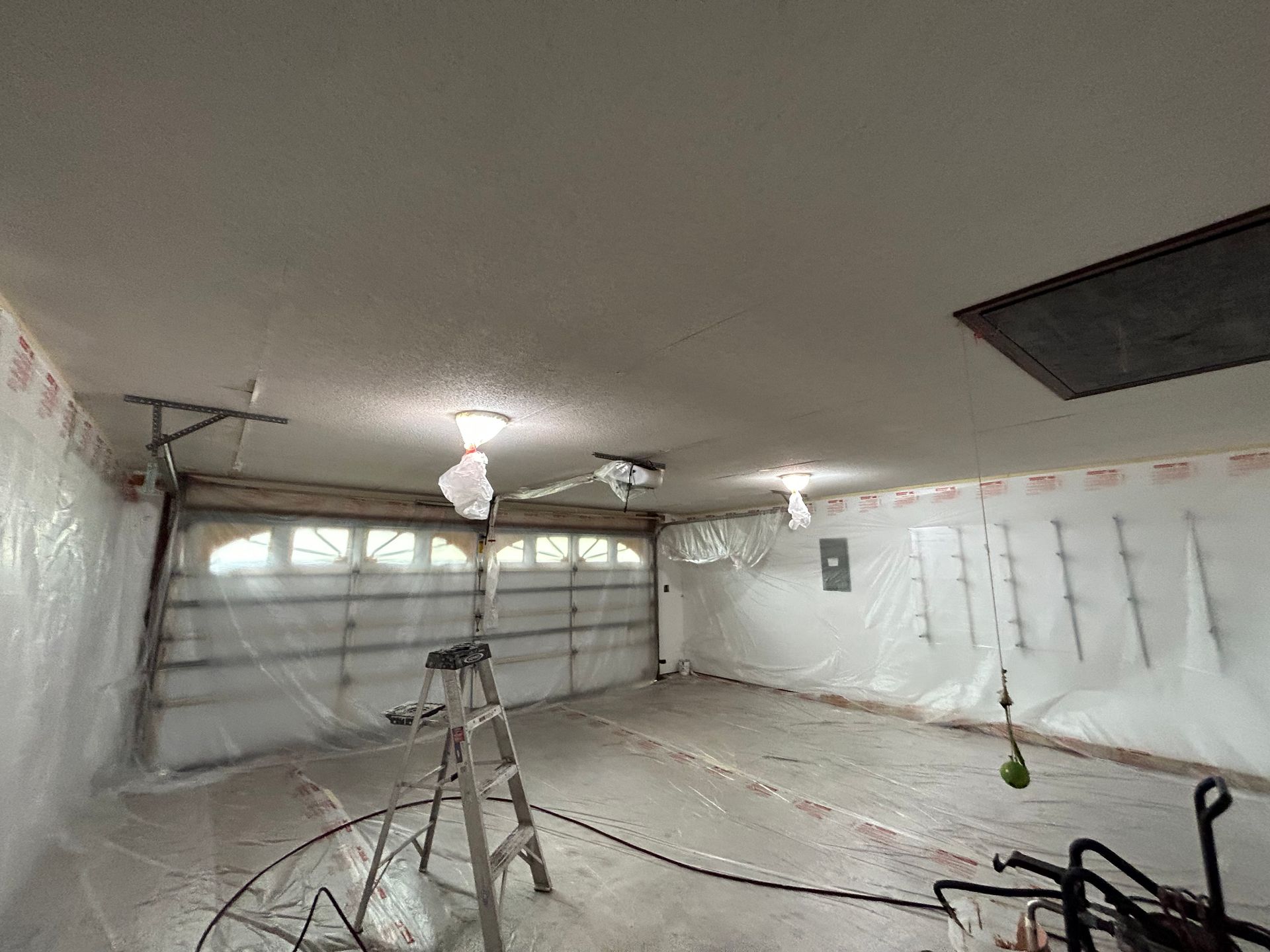 Garage interior during renovation; plastic sheeting protects walls. Garage door closed, ladder in center.
