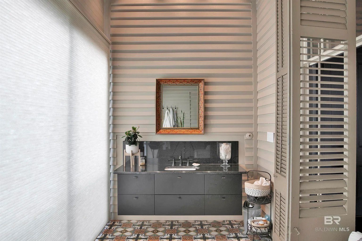 Powder room with gray vanity, gold mirror, patterned tile, and louvered shutters.
