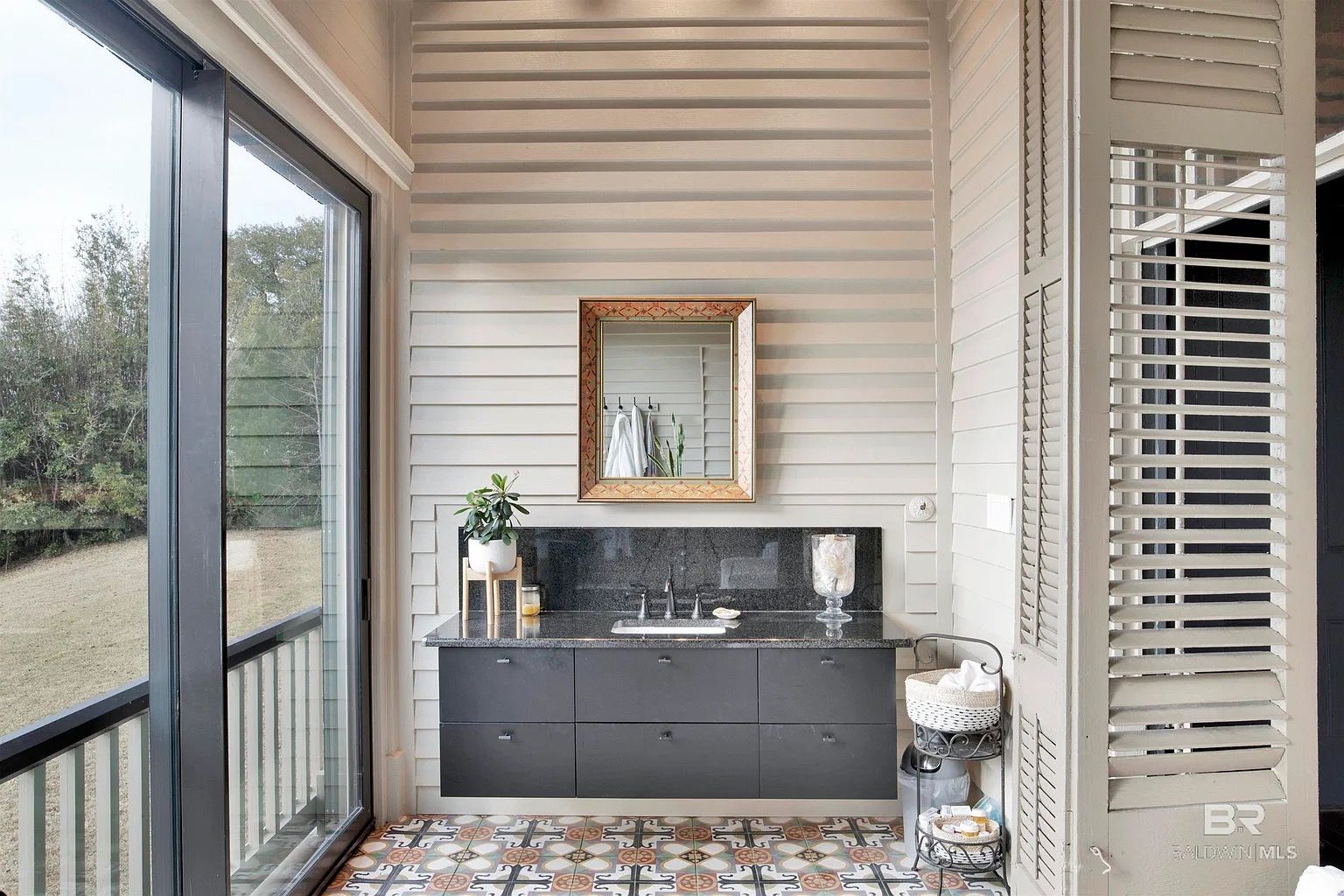 Bathroom with dark countertop, patterned floor, window overlooking greenery, and shuttered wall.