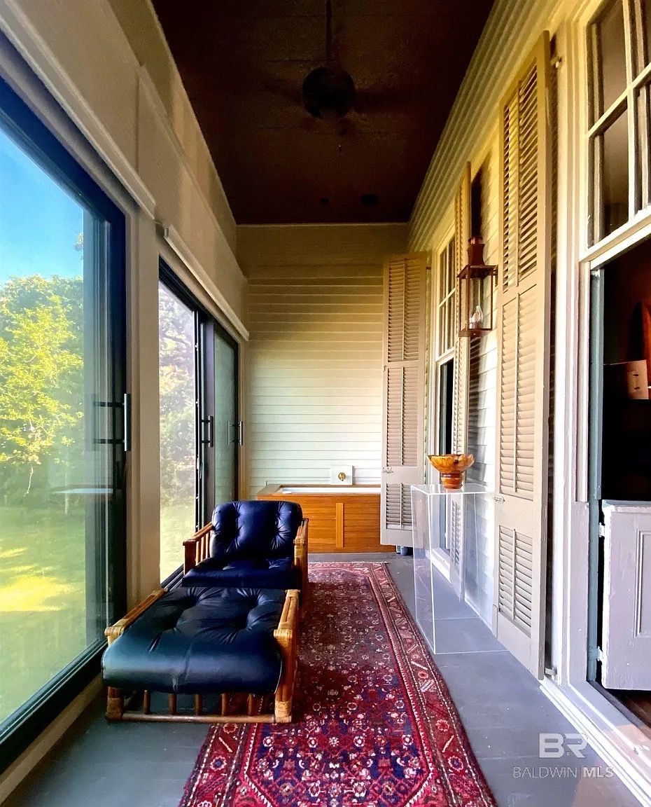 Sunroom with chaise lounge, red rug, and window shutters; view of outdoors.