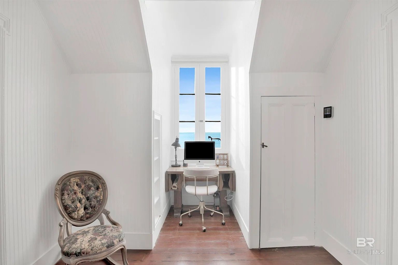 A narrow, white home office nook with a desk facing a window overlooking a blue ocean.