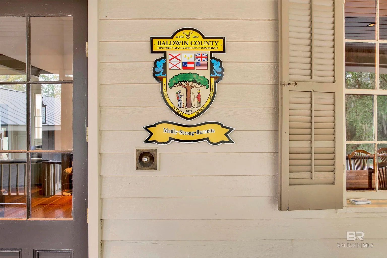 Seal of Baldwin County, Alabama on a white clapboard wall, next to a window with open shutters.