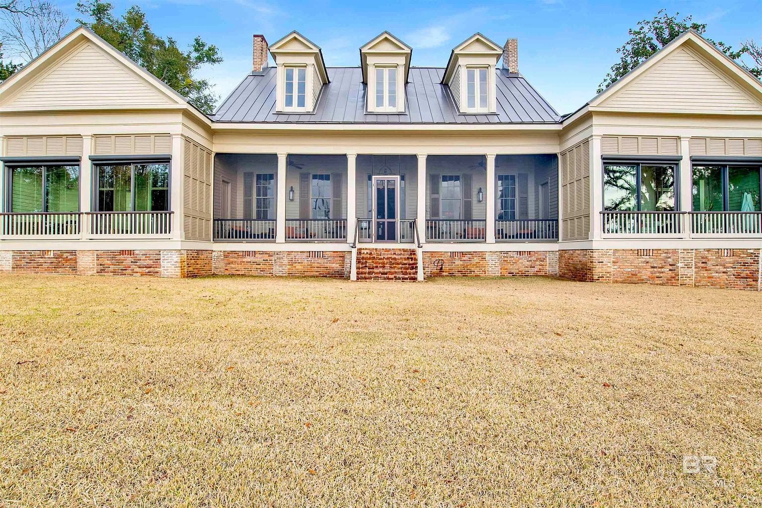 Elegant, long, light-colored house with a screened porch, three dormers, and brick foundation set on a gravel yard.