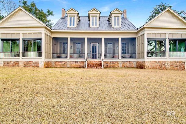 Front view of a large, light-colored house with a wraparound porch, brick foundation, and a gravel yard.