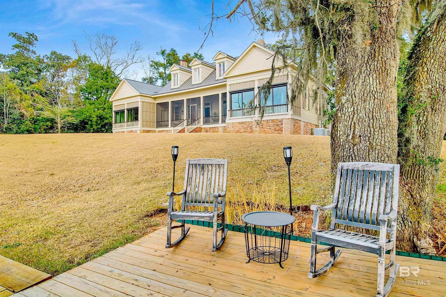 Two weathered rocking chairs and table on a wooden dock, overlooking a house on a grassy hill.