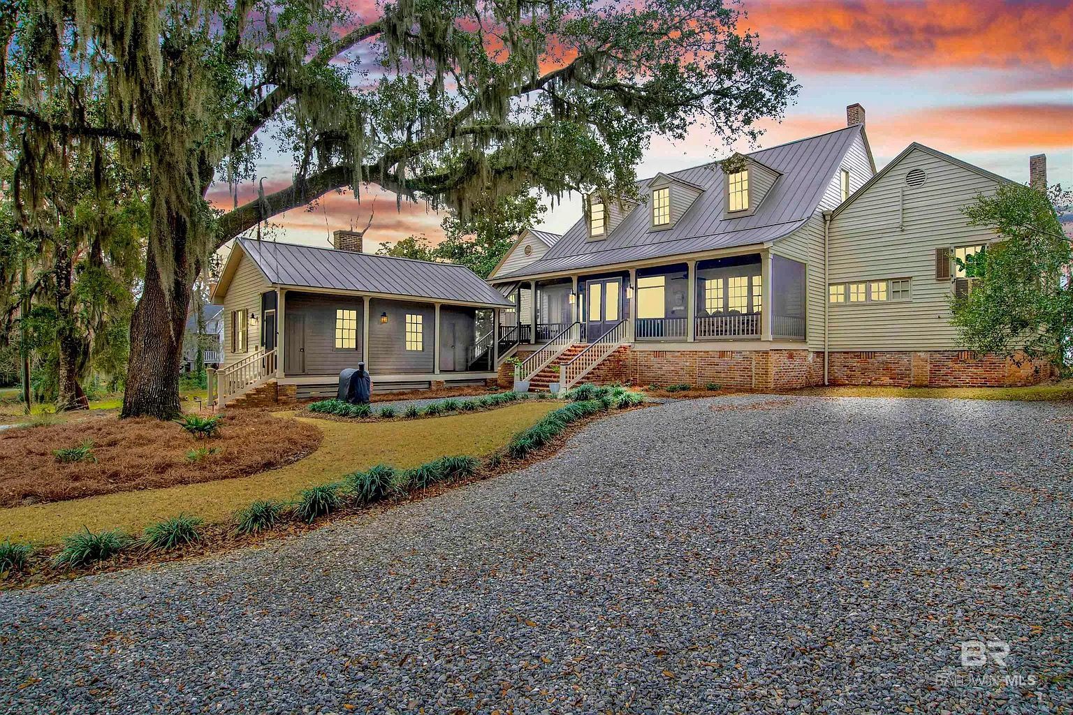 A house with a gravel driveway, lush landscaping, and a sunset sky.