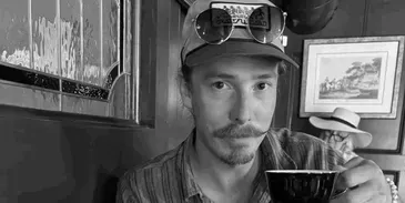 Man with mustache, hat, sunglasses, and cup, in a dimly lit room with framed pictures.