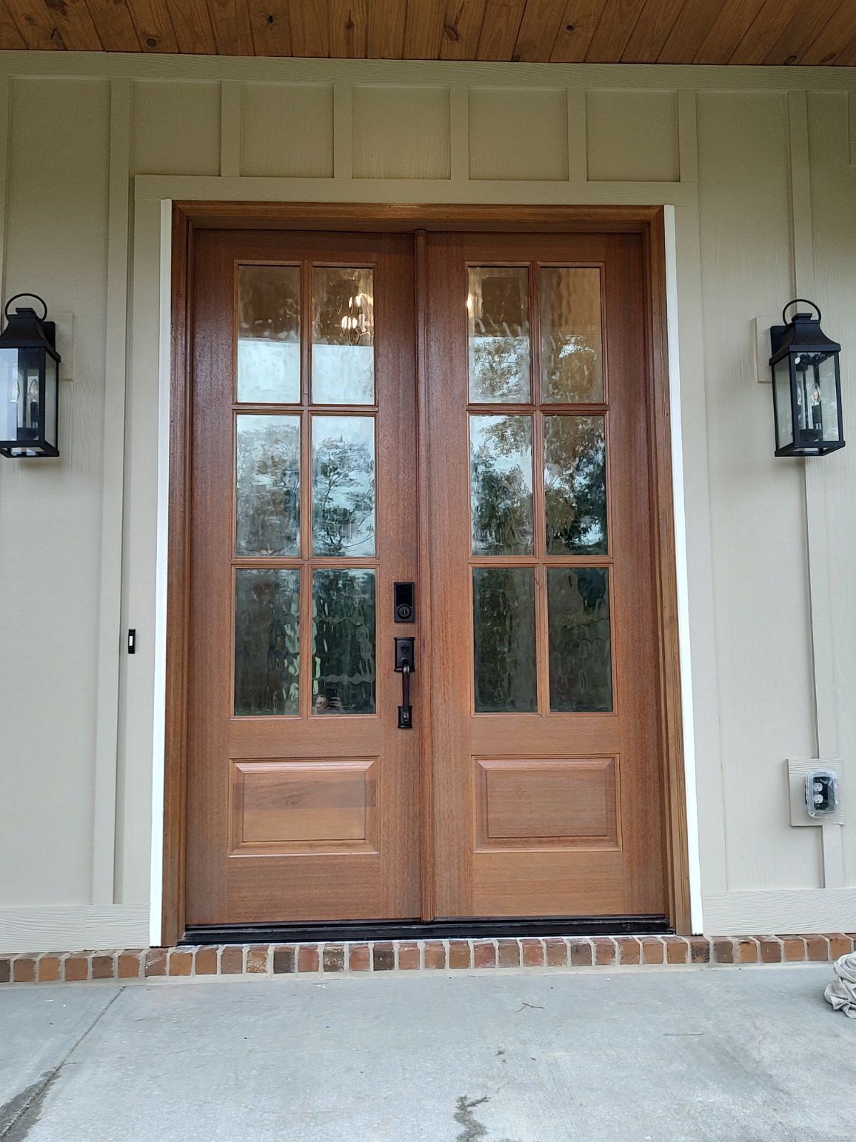 Double wooden front doors with glass panes and black hardware, flanked by lanterns on a beige exterior.
