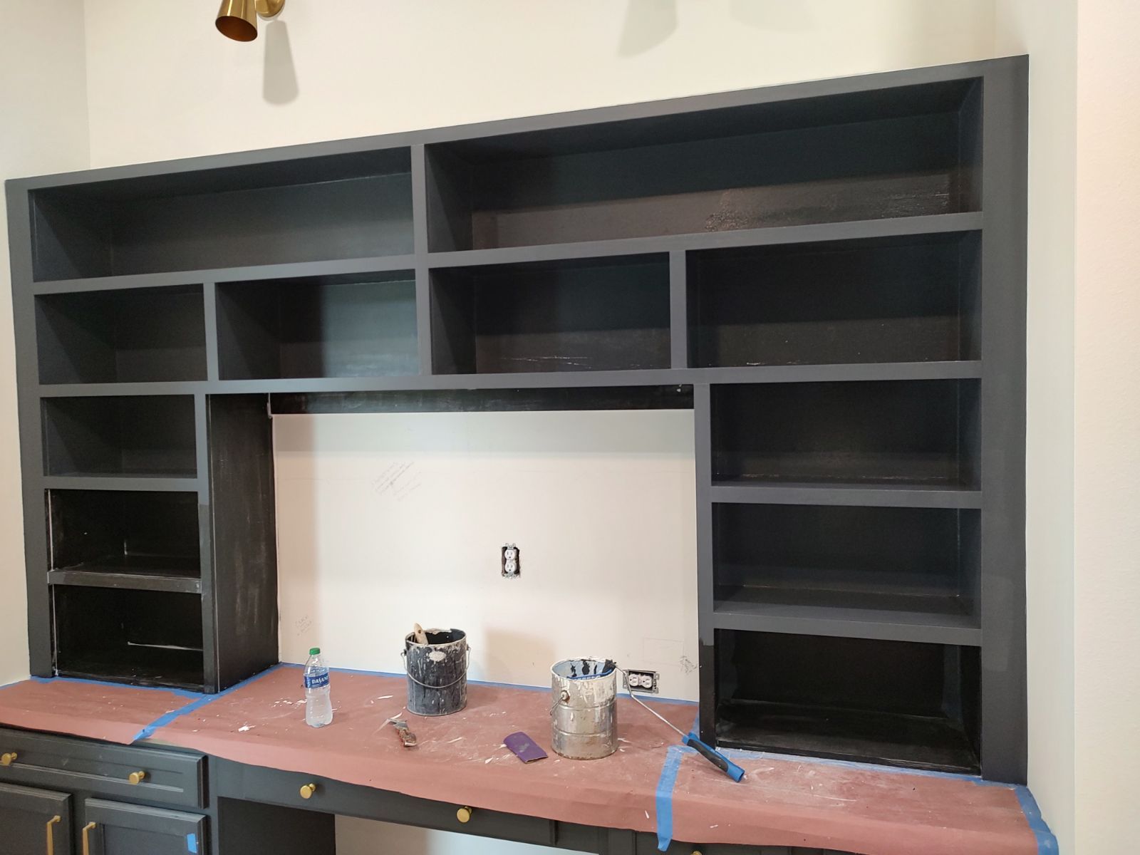 Dark gray built-in desk with bookshelves. Empty center space. Painting in progress; a room interior.