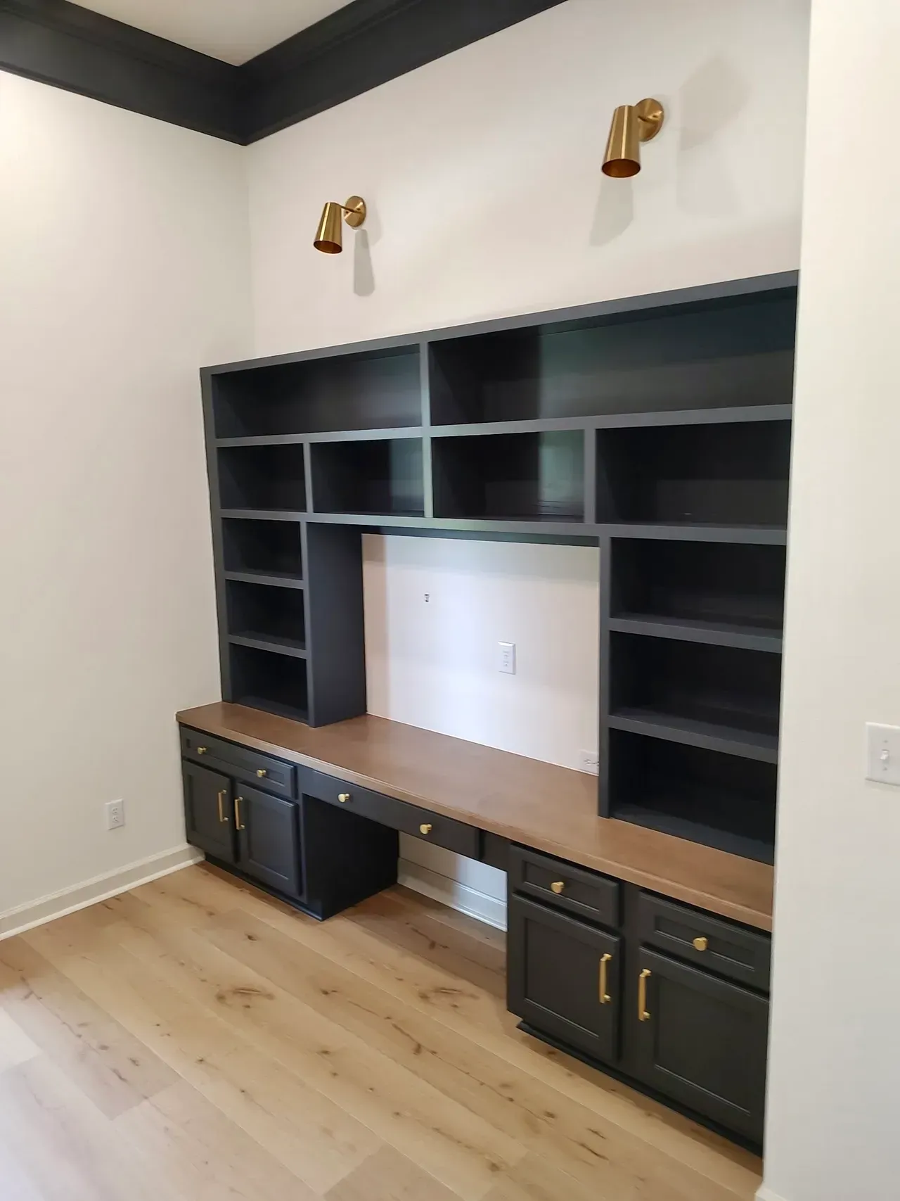 Dark gray built-in desk and shelves with gold hardware against white walls. Two gold sconces.