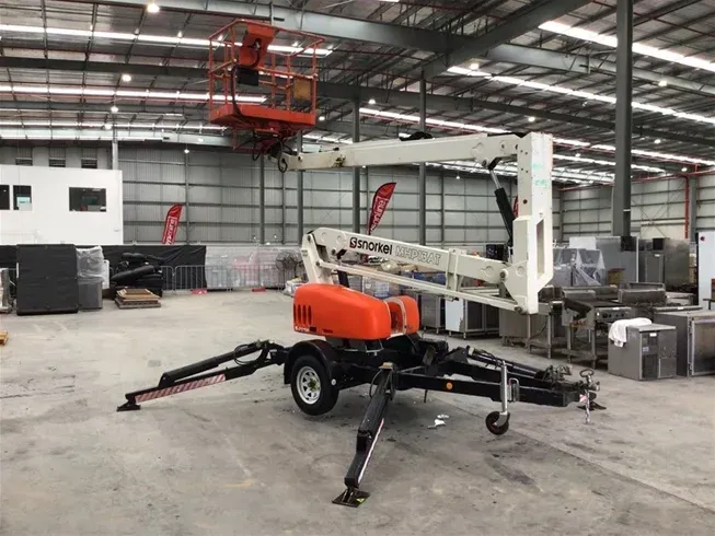 A white and orange trailer-mounted boom lift with stabilizers deployed, standing in a large indoor warehouse — Superior Access Hire in Wynnum, QLD