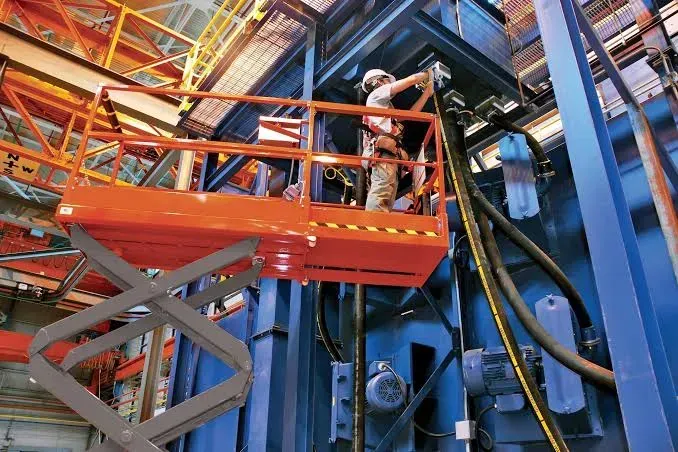 A worker in a safety harness stands on a raised orange scissor lift, performing maintenance on a large blue industrial unit — Superior Access Hire in Wynnum, QLD