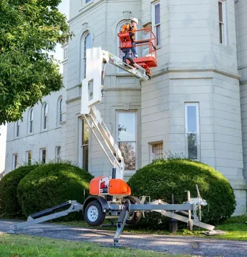 A worker in a safety harness stands in a cherry picker lift, repairing a window on the side of a tall white stone building — Superior Access Hire in Wynnum, QLD