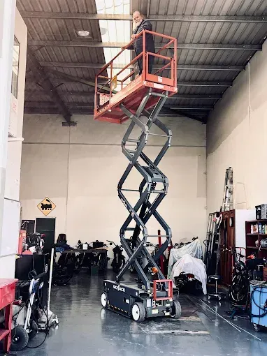 A person stands on a fully extended red scissor lift inside a warehouse or garage, reaching toward the ceiling — Superior Access Hire in Wynnum, QLD