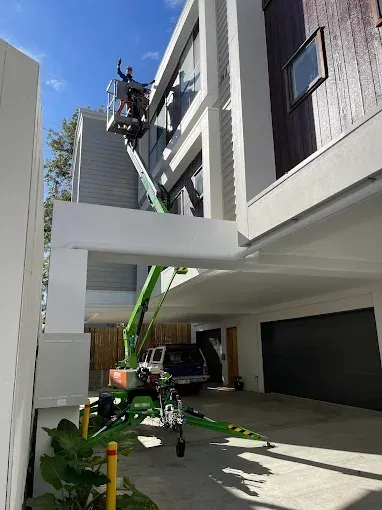 A worker in a bucket lift repairs the exterior of a white multi-story modern building under a clear blue sky — Superior Access Hire in Wynnum, QLD