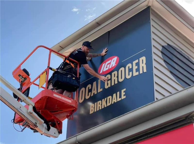 Man in a lift installing an IGA Local Grocer sign in Birkdale.