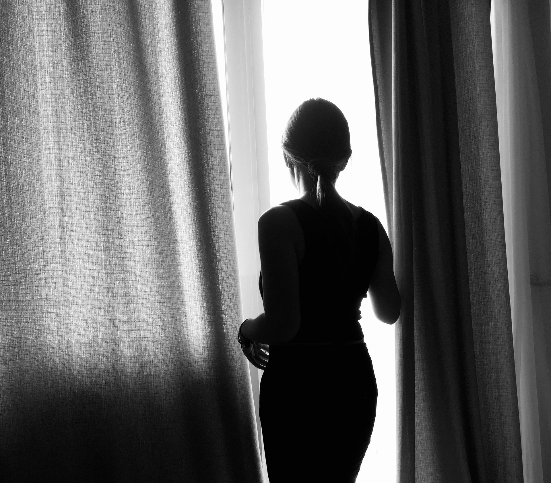 A black-and-white silhouette of a person standing between two curtains, facing a bright, light-filled window.