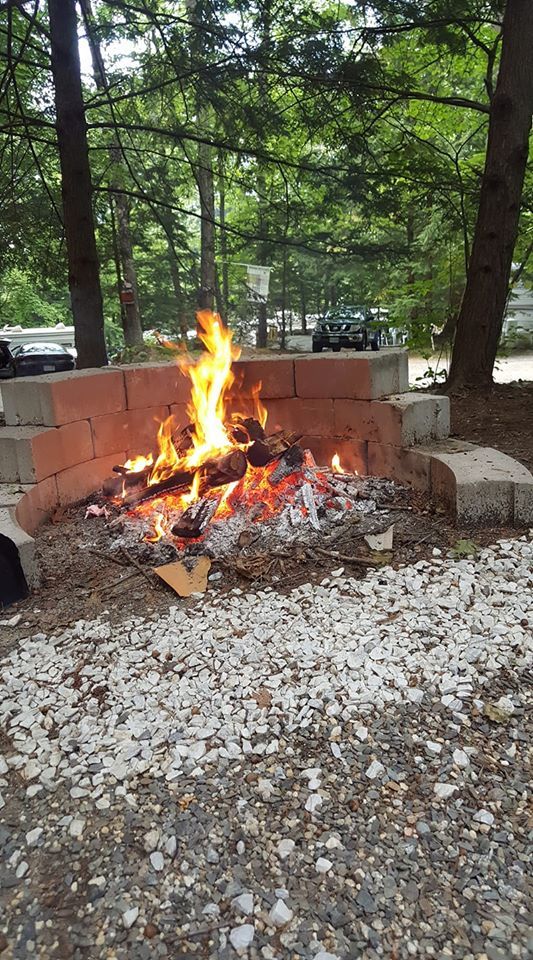 A fire burns in a brick enclosure. Smoke rises, trees in the background, gray gravel ground.