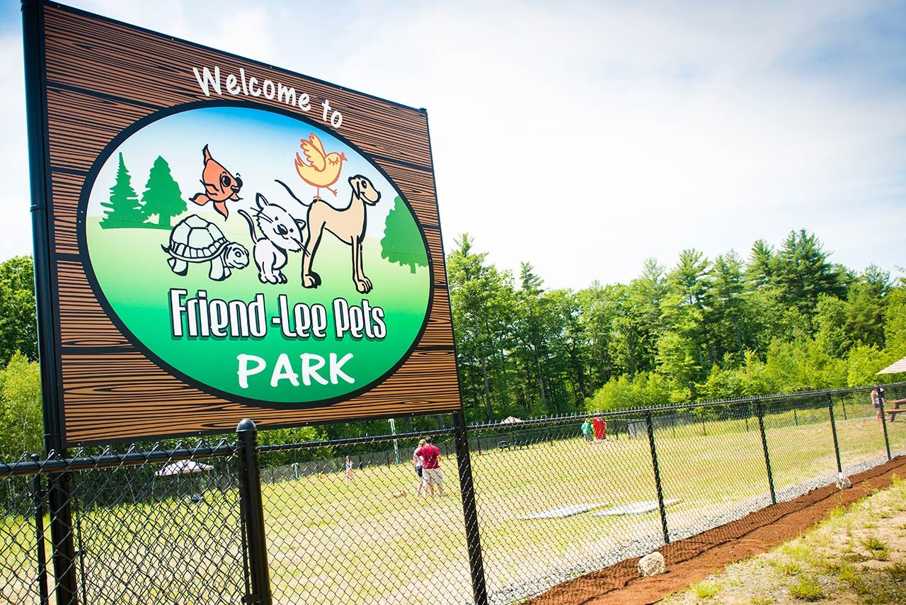 Sign for Friend-two-Pets Park with cartoon animals, fenced dog park, people in distance, green trees, and blue sky.