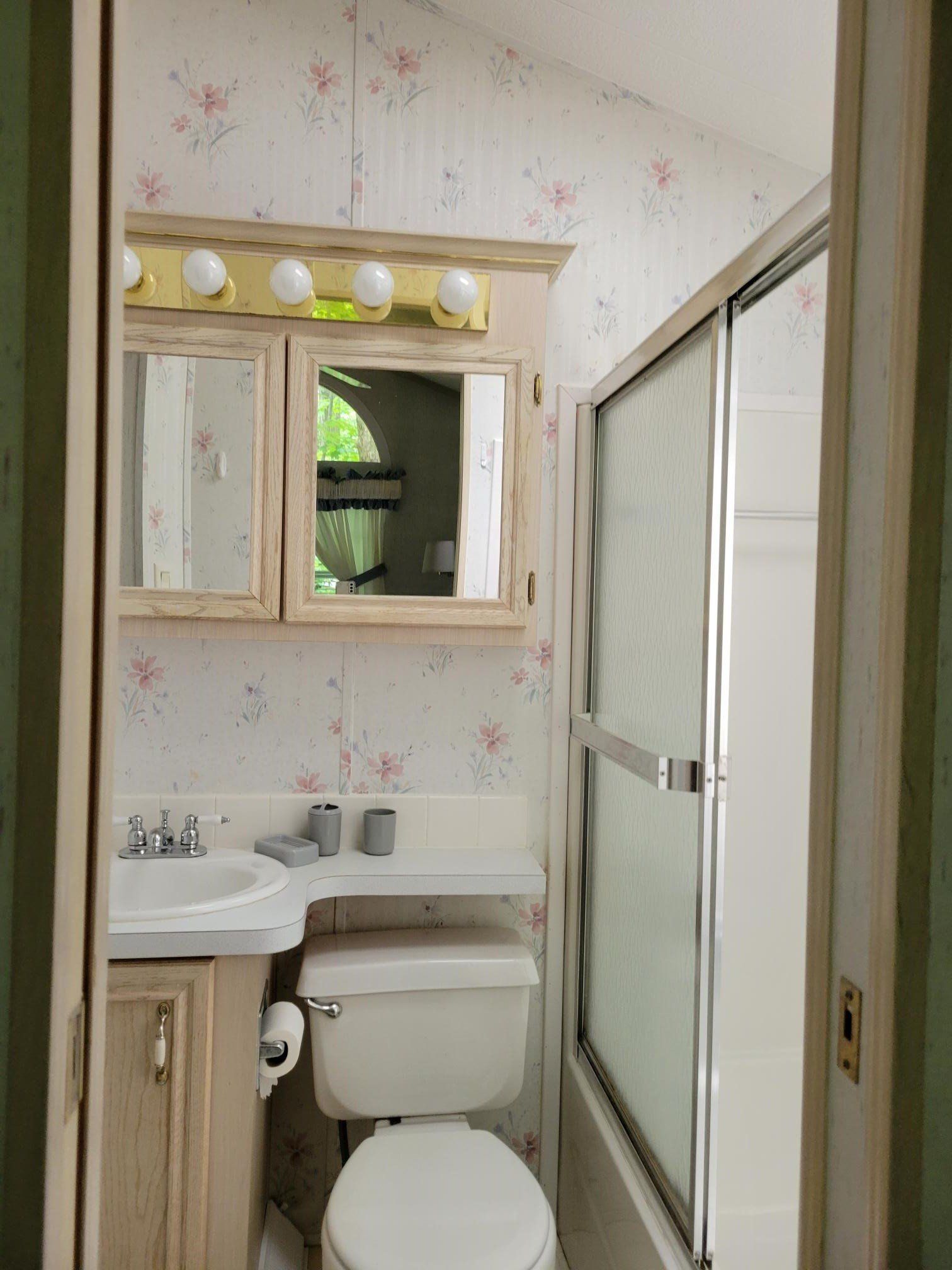 Small bathroom with floral wallpaper, toilet, sink, medicine cabinet, and shower with sliding glass door.