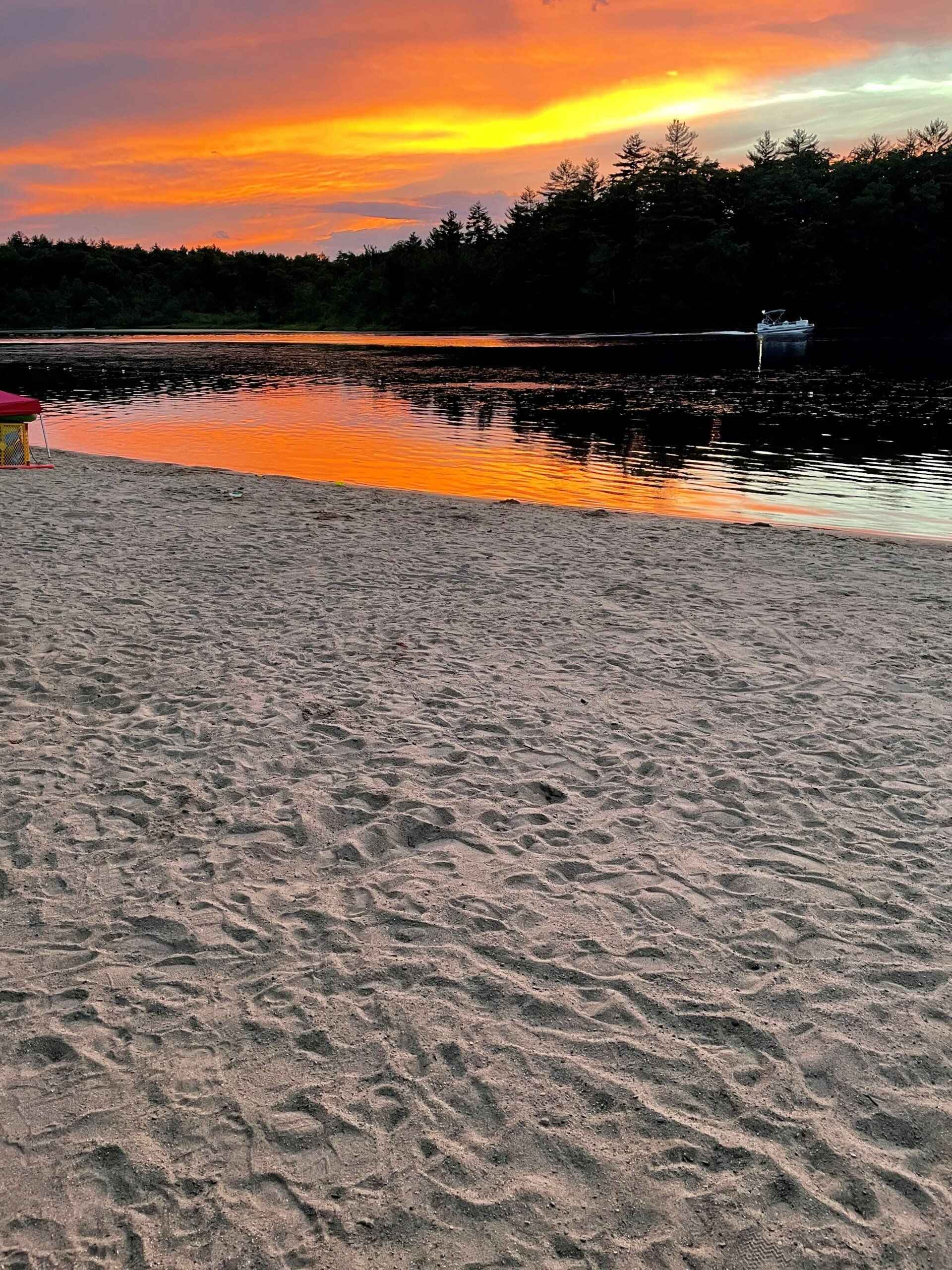 Beach scene at sunset. Orange and yellow sky reflects on water. Sand in foreground. Trees in background.