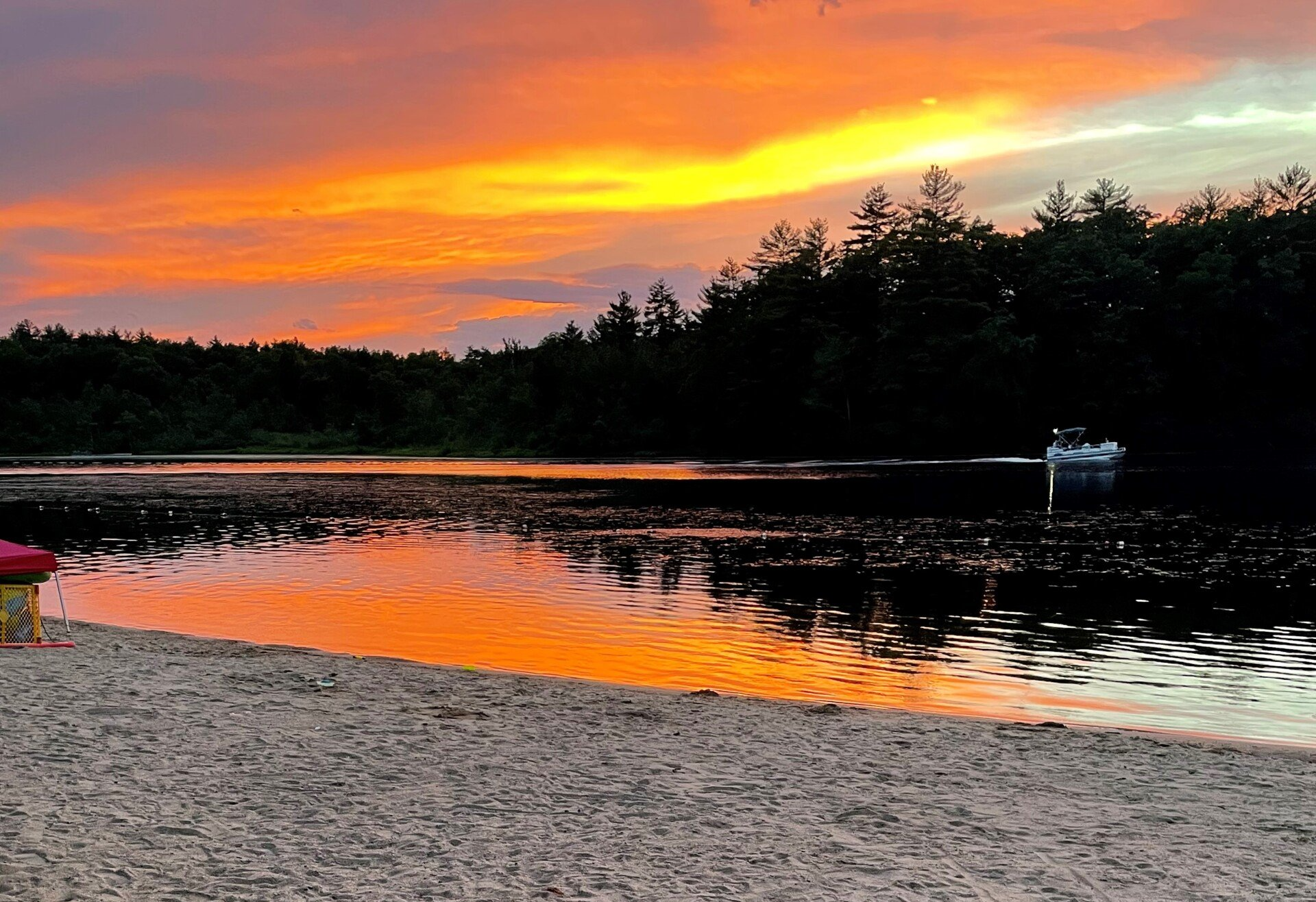 Sunset over a lake; orange and yellow sky reflects in water. Boat floats on lake, trees in silhouette on horizon.