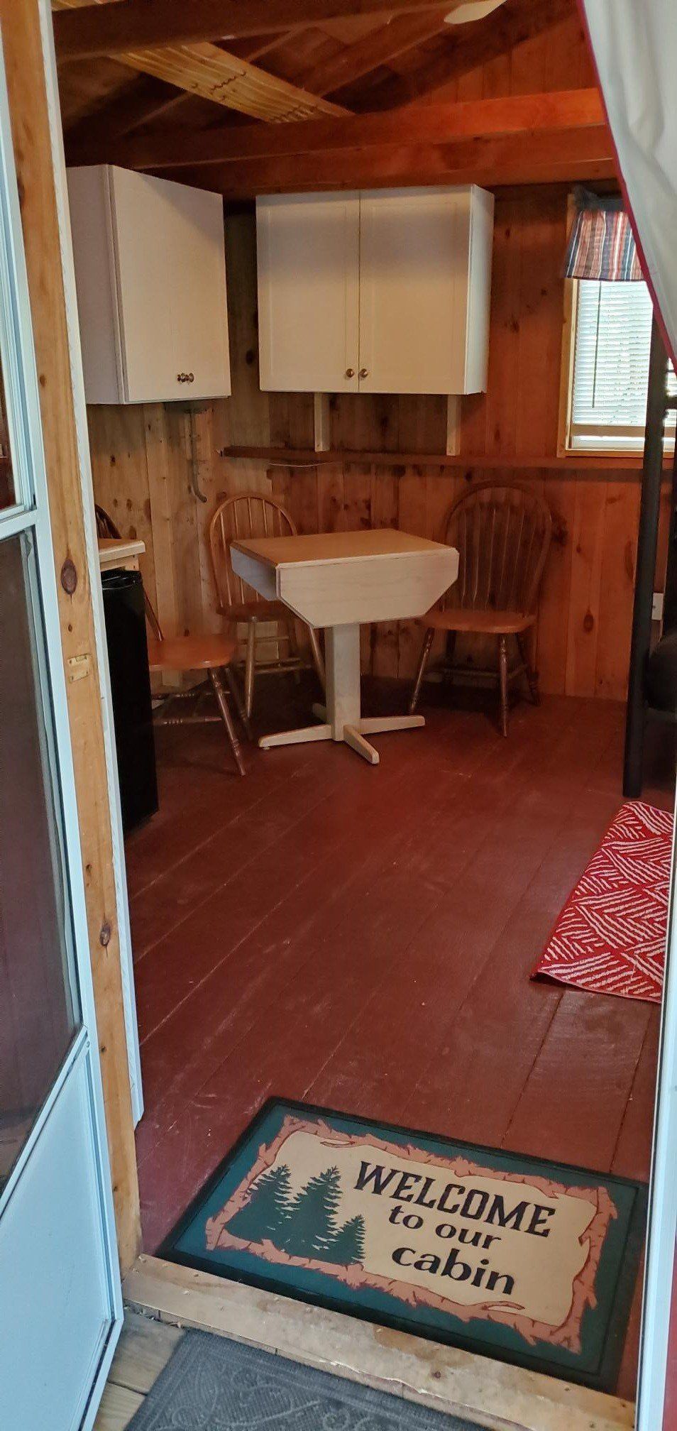 Interior of a cabin with white cabinets, a table and chairs, and a welcome mat. Red floors and wood walls.