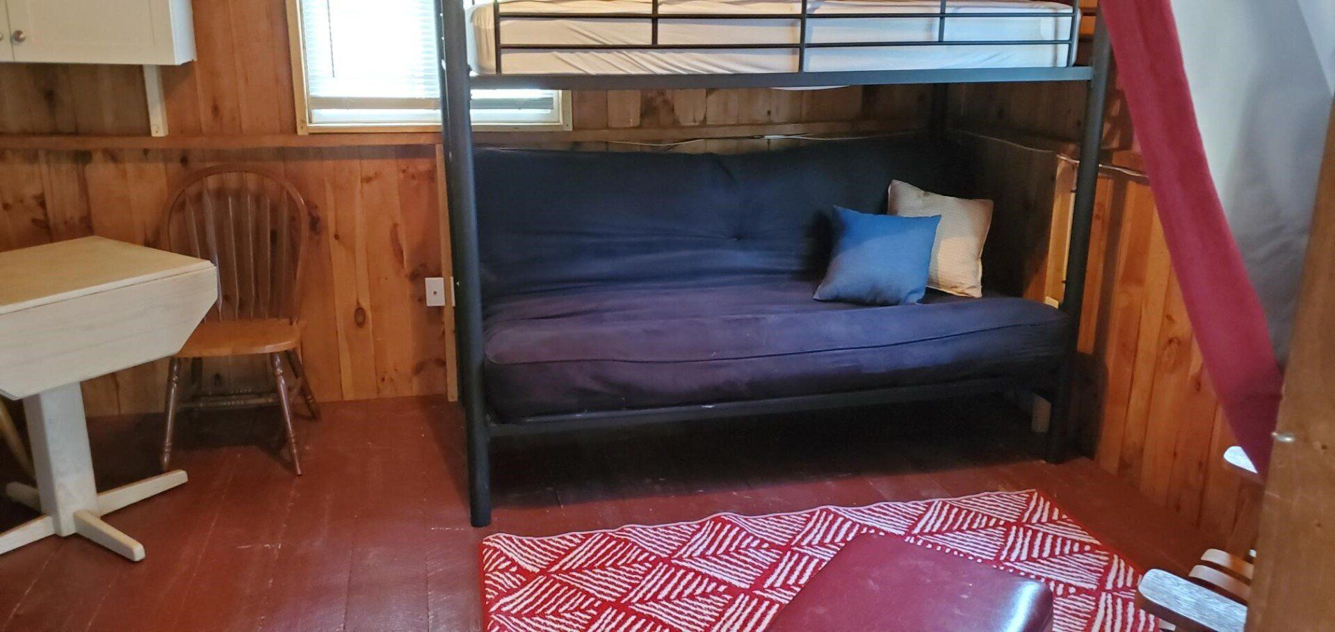 Room with bunk bed over a futon, drop-leaf table, chair, and rug with red pattern. Wood paneling on the walls.