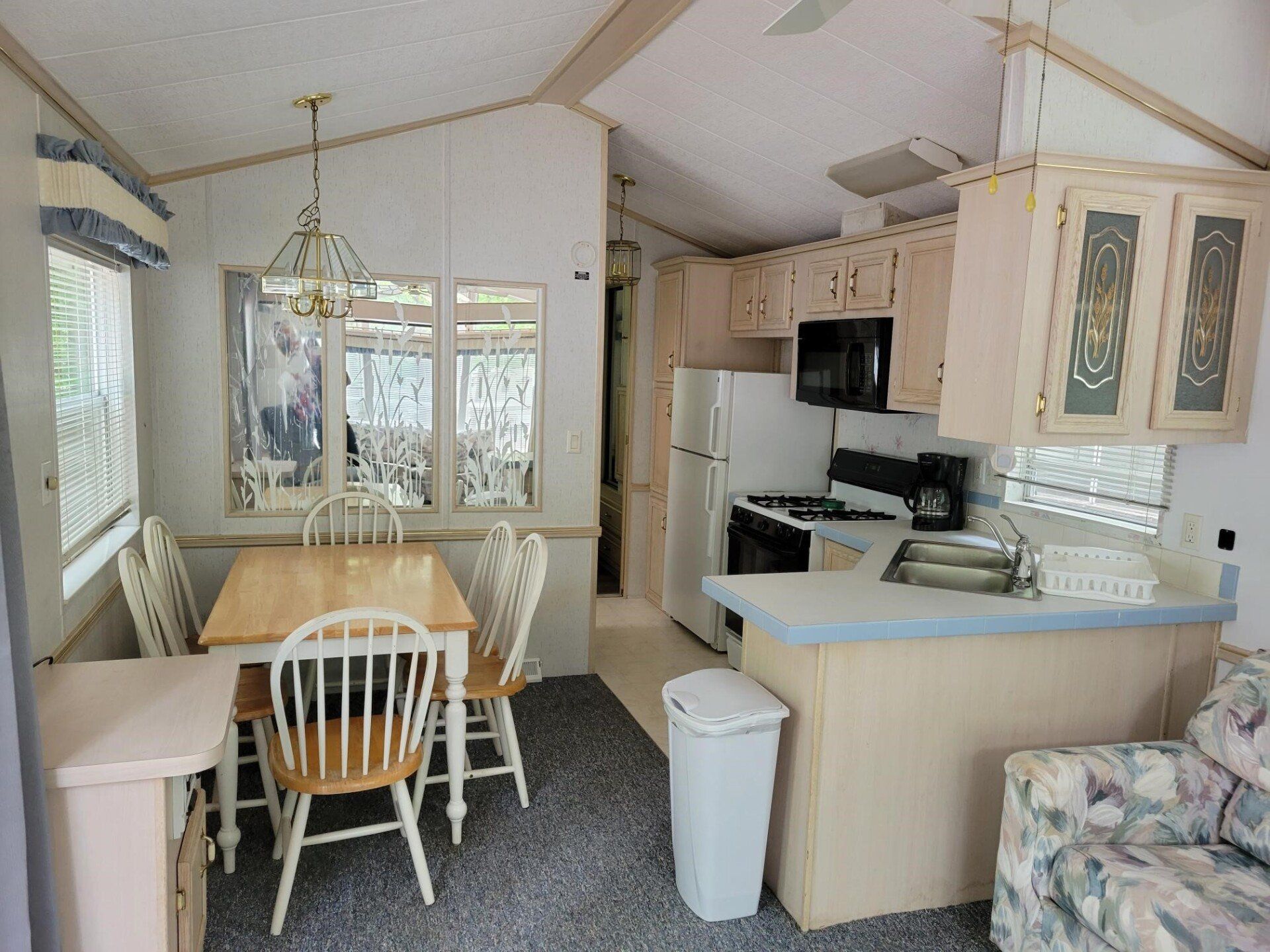 Interior of a mobile home: kitchen and dining area. Light-colored cabinets, appliances, table and chairs, and a small sofa.
