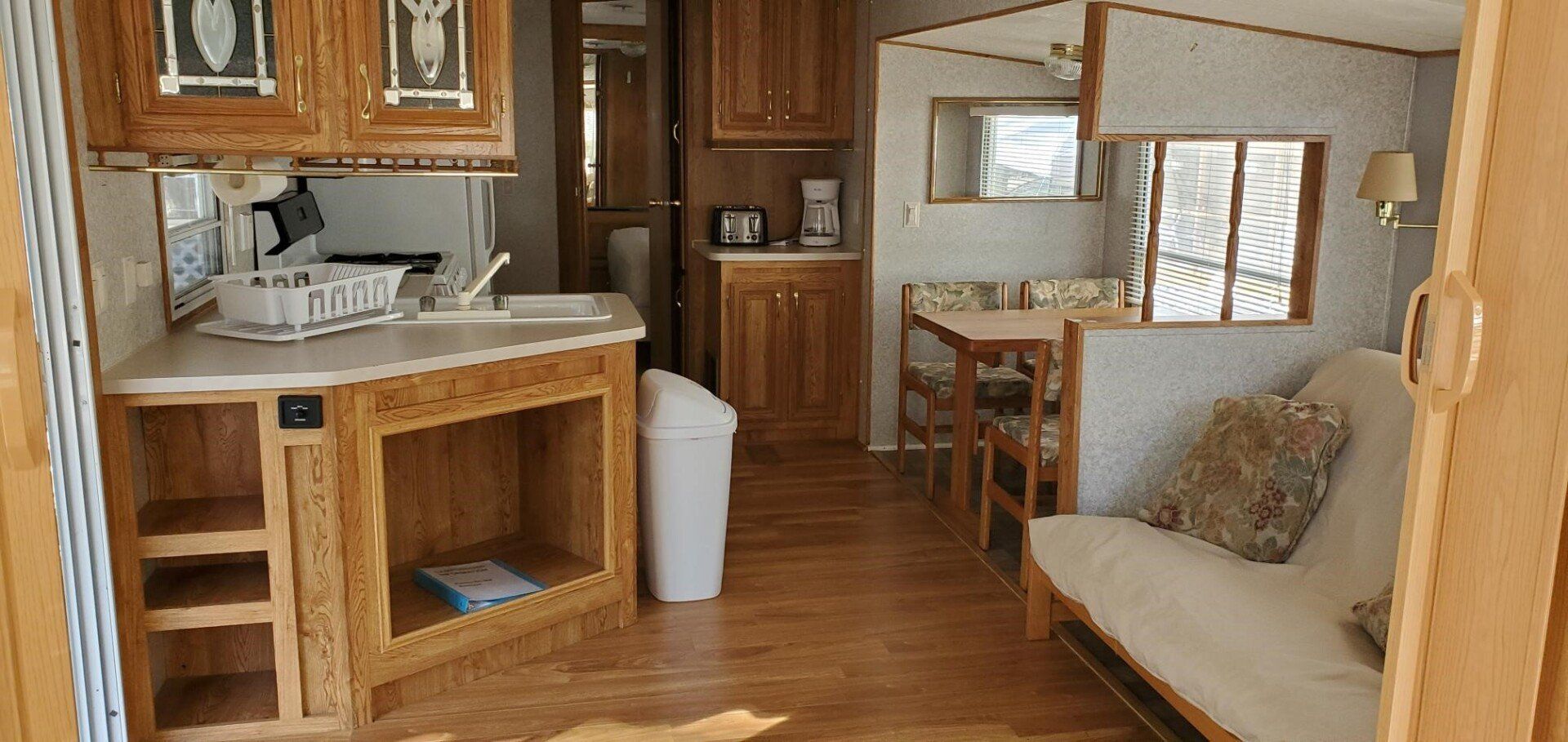 Interior of a small cabin or mobile home with a kitchen, dining area, and futon. Wooden cabinets and flooring.