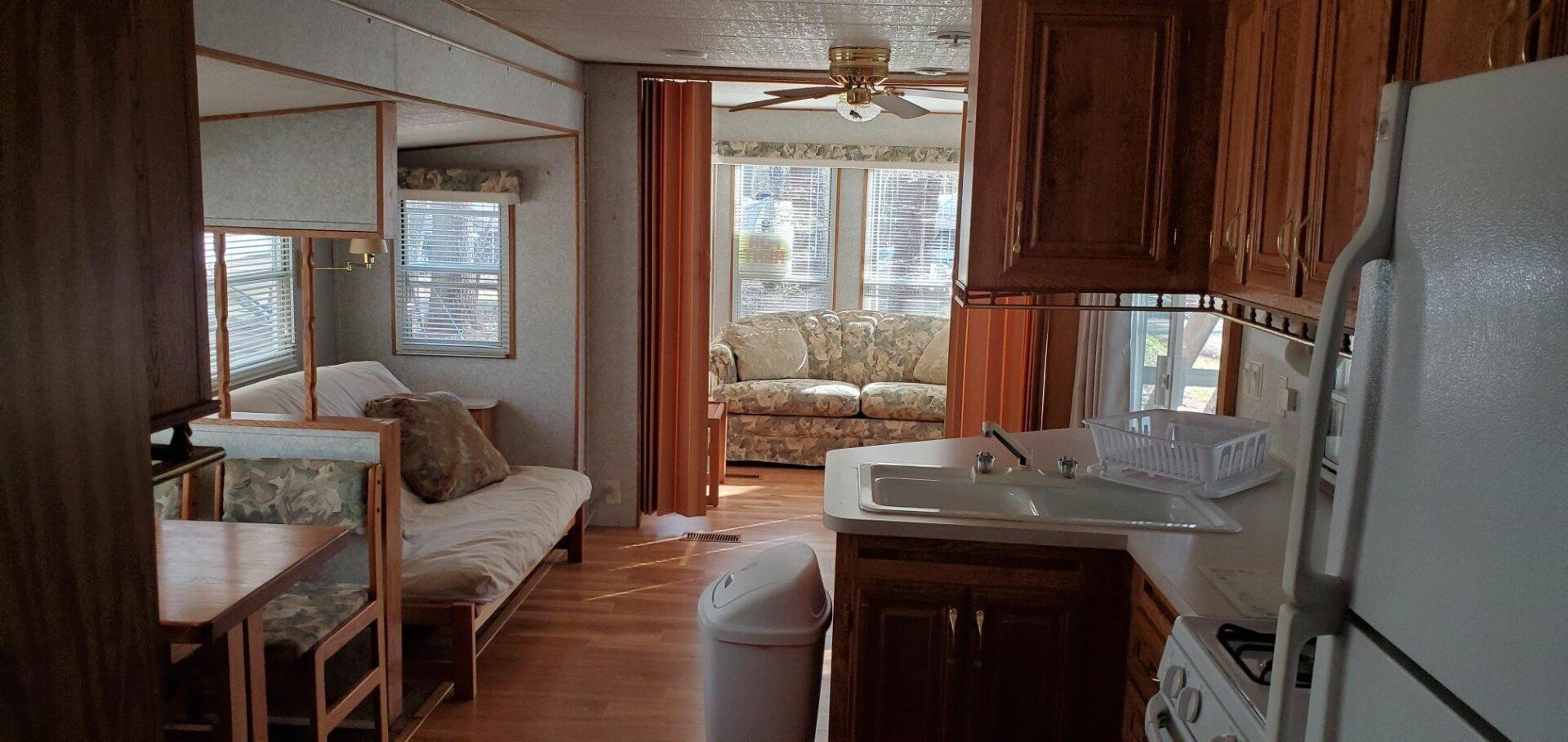Interior of a trailer, with kitchen, living room, and a futon, wooden cabinets.