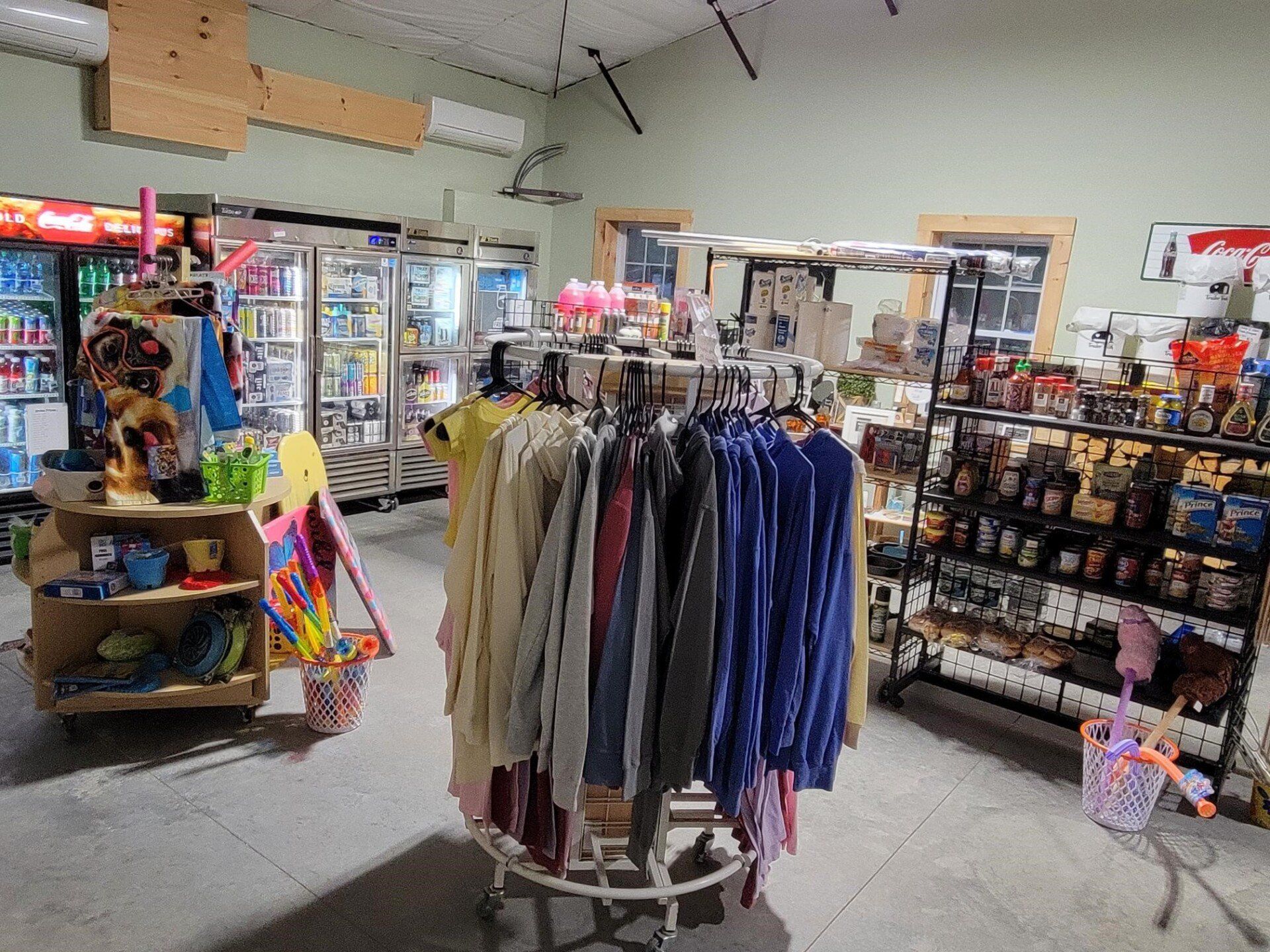 Inside a store with clothes rack, shelves of goods, and refrigerated drinks.