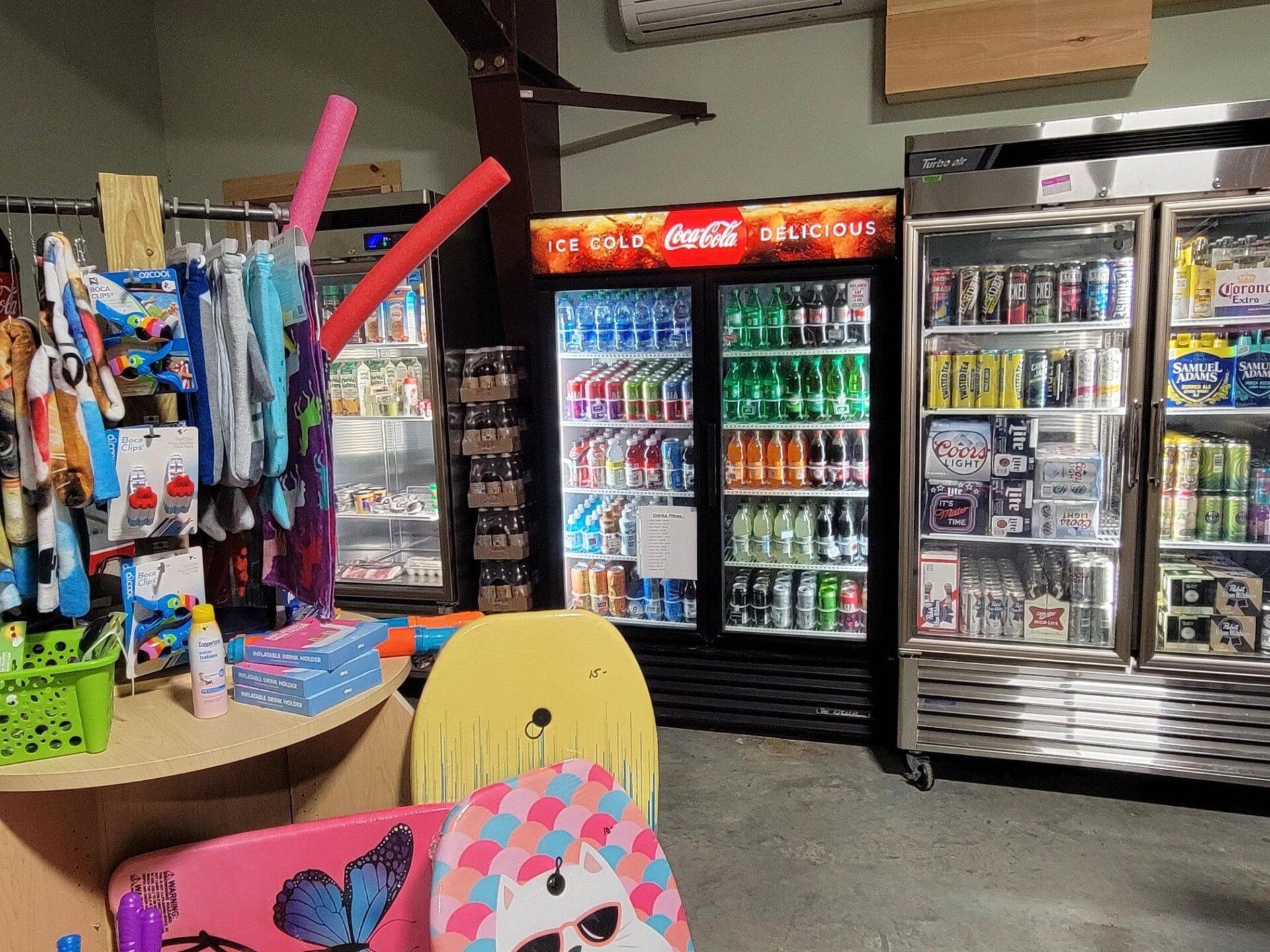 Inside a shop, refrigerators with drinks, surfboards, clothing, and other merchandise are visible.