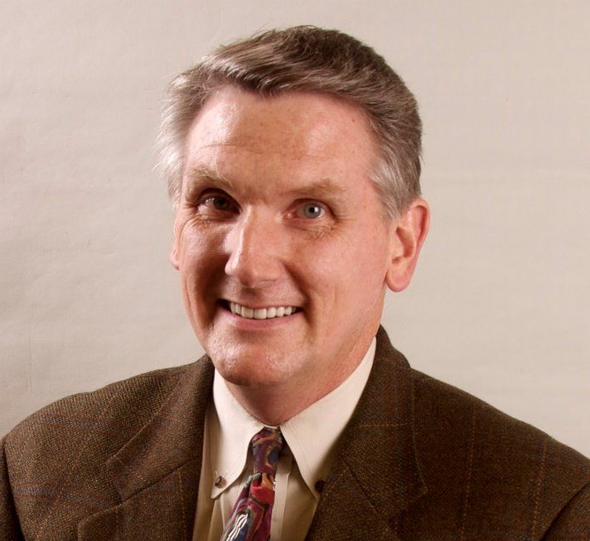 Smiling man in a brown suit and patterned tie against a plain background