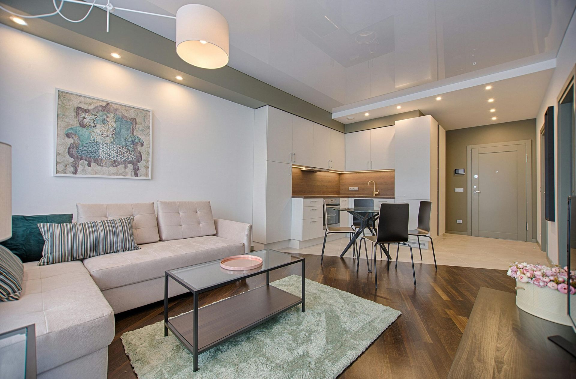 Modern open-plan living room with beige sectional, glass coffee table, dining area, and kitchen in the background