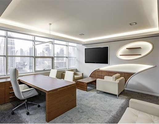 Modern office with wooden desk, white chairs, gray carpet, sofa, wall-mounted TV, and large windows.