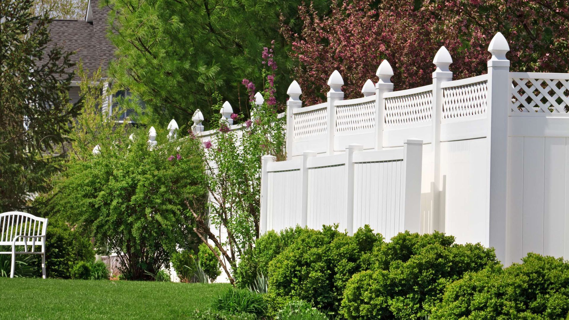 A white fence surrounds a lush green yard with trees and bushes.