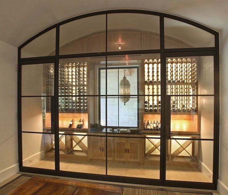 Wine cellar behind a black-framed glass wall with arched top. Includes wooden shelves, wine bottles, and a small table.