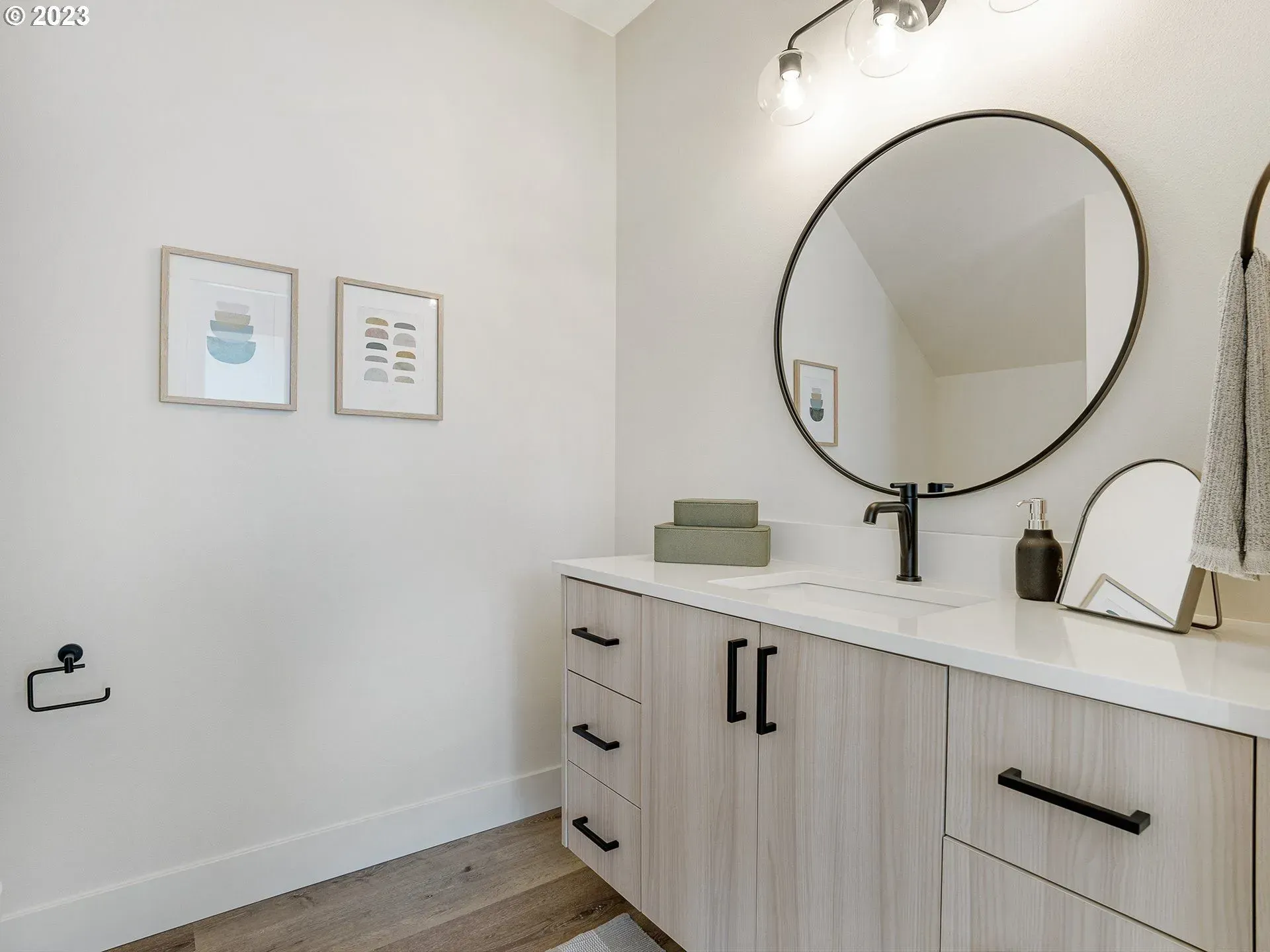 Bathroom with light wood vanity, round mirror, black fixtures, and two framed prints.